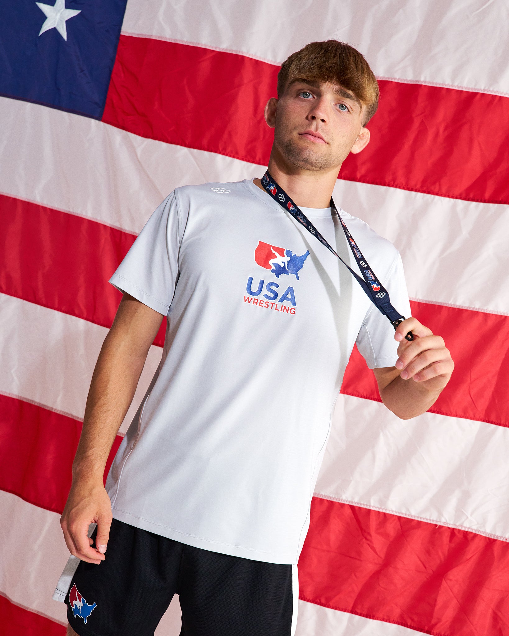 A young man wearing a white USA Wrestling t-shirt and black athletic shorts stands in front of a large American flag backdrop, holding a medal around his neck.
