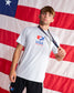 A young man wearing a white USA Wrestling t-shirt and black athletic shorts stands in front of a large American flag backdrop, holding a medal around his neck.