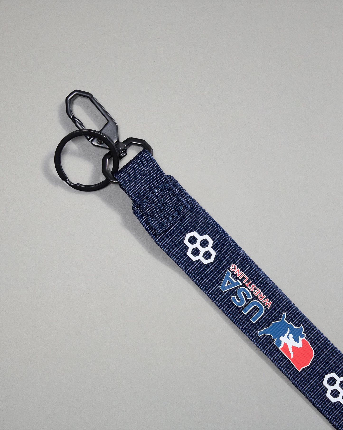 Close-up of the navy USA Wrestling lanyard featuring a durable black metal key clip, key ring, woven polyester strap, USA Wrestling logo, and RUDIS honeycomb branding.