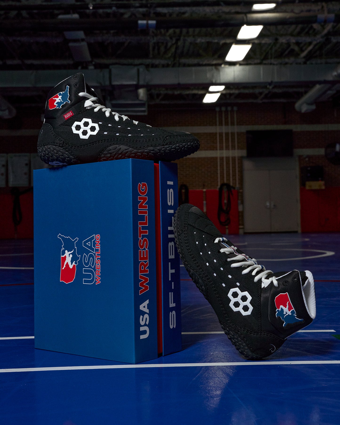 Black wrestling shoes with USA Wrestling logo on a blue box in an indoor sports facility.