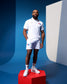 An adult male model wearing a white USA Wrestling athletic polo and shorts, standing on a red pedestal with a blue and white background.