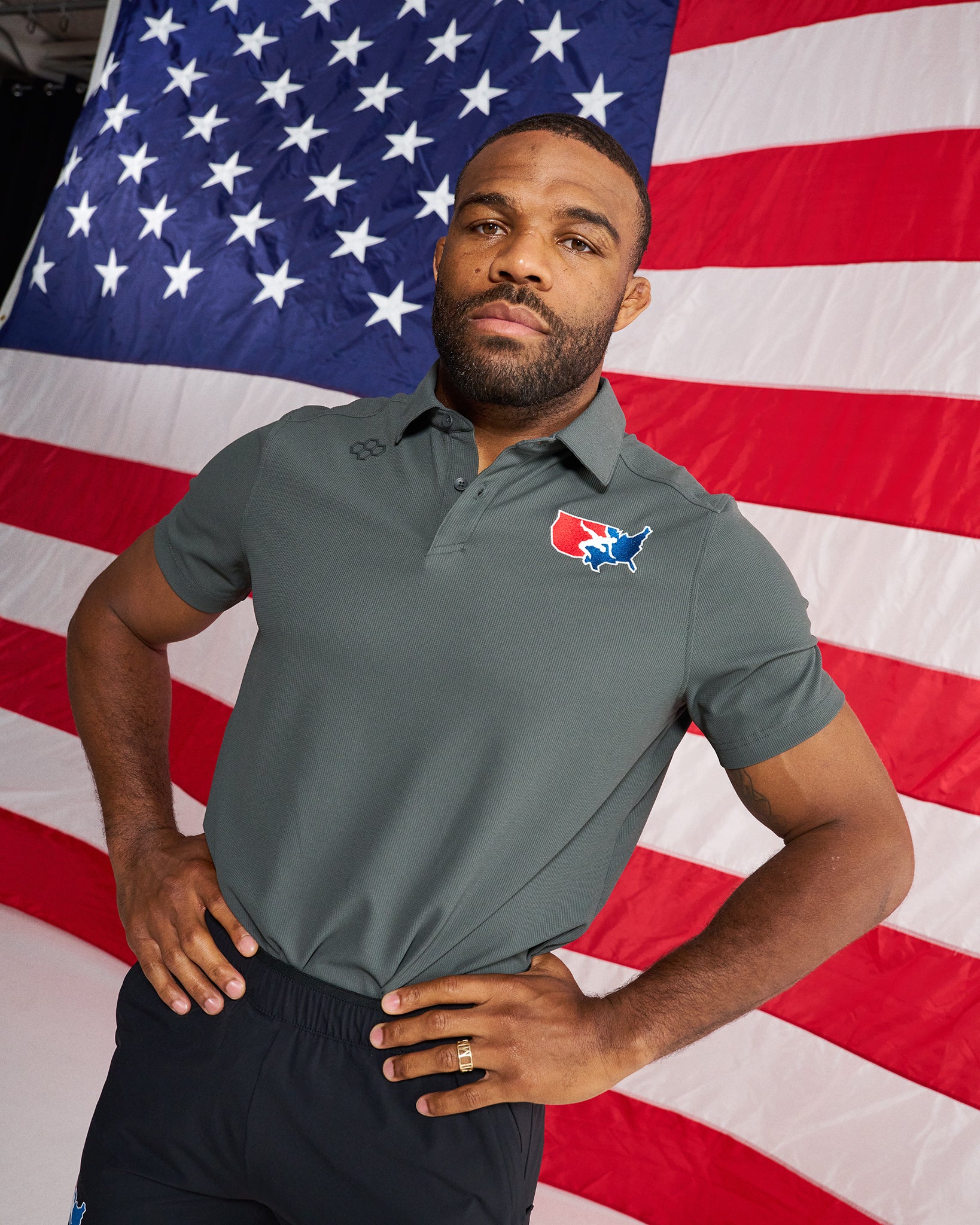 A man wearing a dark gray polo shirt with a red, white, and blue logo on the left chest, posed in front of a large American flag backdrop.
