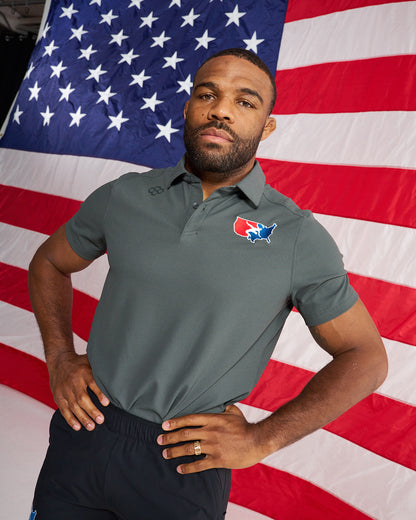 A man wearing a dark gray polo shirt with a red, white, and blue logo on the left chest, posed in front of a large American flag backdrop.