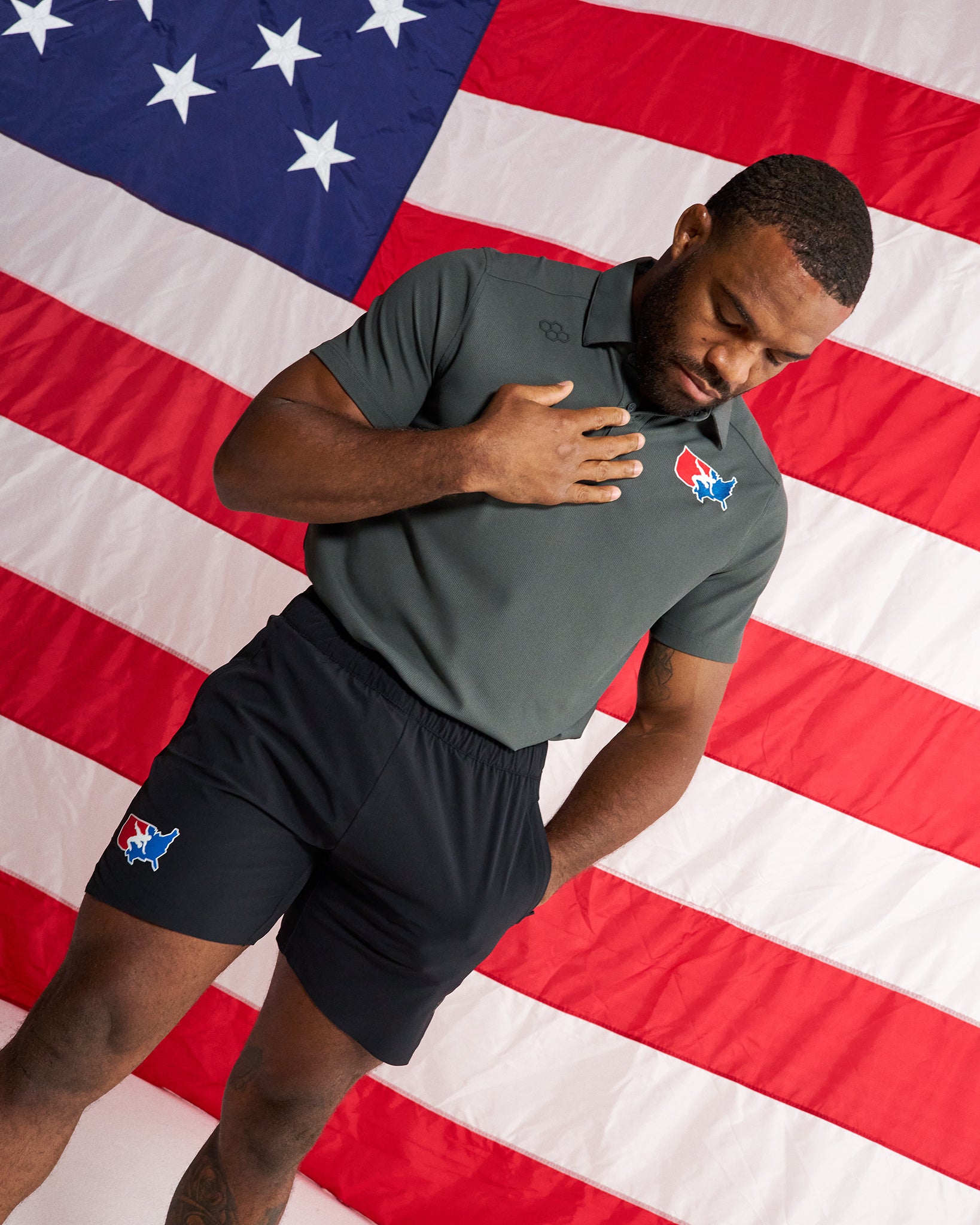 A man wearing a dark gray short-sleeve polo shirt and black athletic shorts stands in front of a large American flag. The shirt has a small red, white, and blue logo on the left chest, and the shorts have a matching logo on the left thigh.