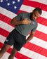 A man wearing a dark gray short-sleeve polo shirt and black athletic shorts stands in front of a large American flag. The shirt has a small red, white, and blue logo on the left chest, and the shorts have a matching logo on the left thigh.
