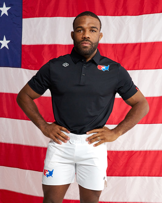 A man stands with hands on hips in front of a large American flag, wearing a black short-sleeve polo shirt with a small logo on the right chest and a red, white, and blue logo on the left chest, paired with white shorts featuring a matching red, white, and blue logo near the left hem.