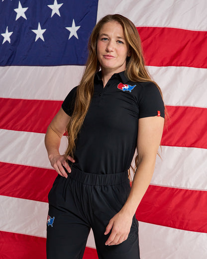 A woman stands in front of a large American flag wearing a black short-sleeve collared athletic top and matching black pants with small red, white, and blue logo patches on the chest and upper thigh.