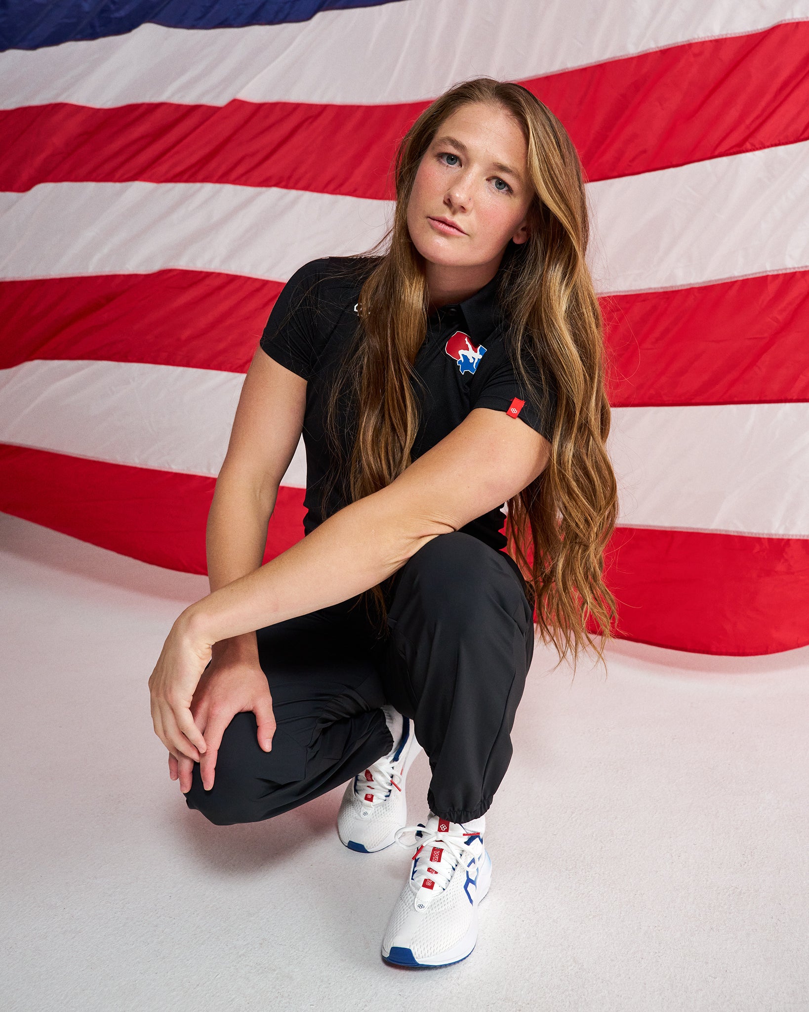 A woman crouches in front of a large American flag backdrop, wearing a black short-sleeve polo shirt with a red, white, and blue logo on the chest, black pants, and white sneakers with red and blue accents.