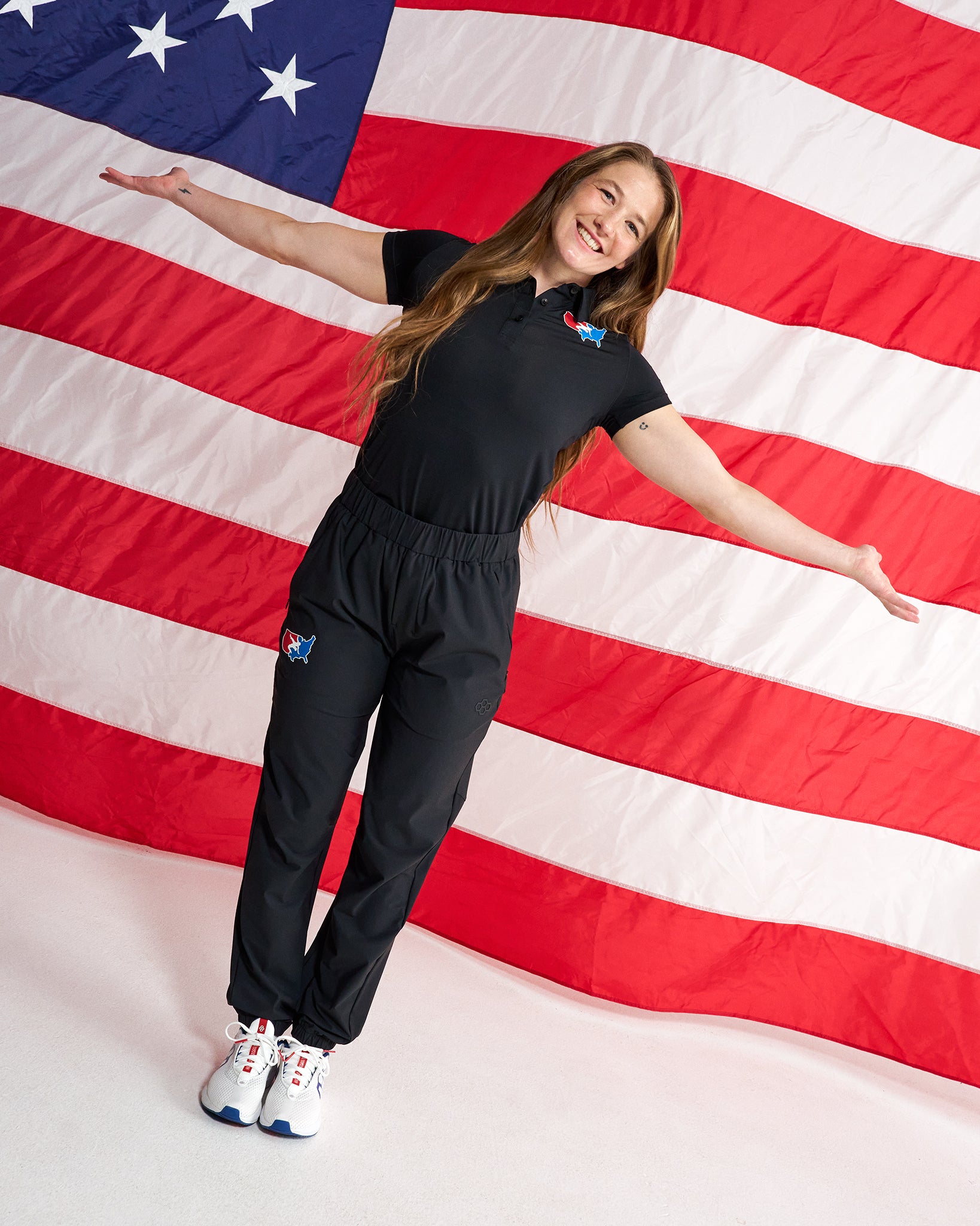A woman wearing a black short-sleeve polo shirt and matching black jogger pants stands with arms outstretched in front of a large American flag backdrop. The outfit has small red, white, and blue logo patches on the chest and left thigh, and she is wearing white sneakers with red and blue accents.