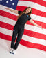A woman wearing a black short-sleeve polo shirt and matching black jogger pants stands with arms outstretched in front of a large American flag backdrop. The outfit has small red, white, and blue logo patches on the chest and left thigh, and she is wearing white sneakers with red and blue accents.