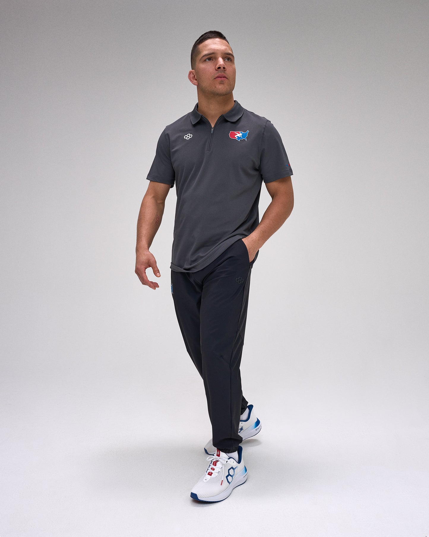 Man wearing a dark gray polo shirt with a 'USA Wrestling' logo and dark pants on a light gray background
