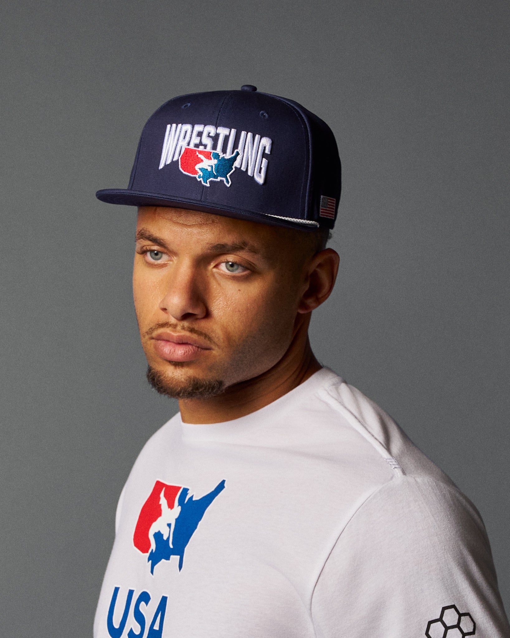 Man wearing a navy 'Wrestling USA' cap and white shirt with logo on a gray background