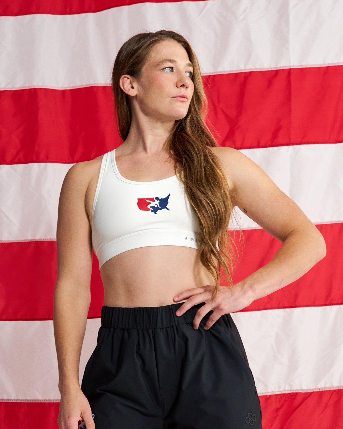 A woman wearing a white sports bra with a red, white, and blue Texas state map graphic on the chest, paired with black athletic shorts, standing in front of a red and white American flag backdrop.