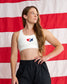 A woman wearing a white sports bra with a red, white, and blue Texas state map graphic on the chest, paired with black athletic shorts, standing in front of a red and white American flag backdrop.