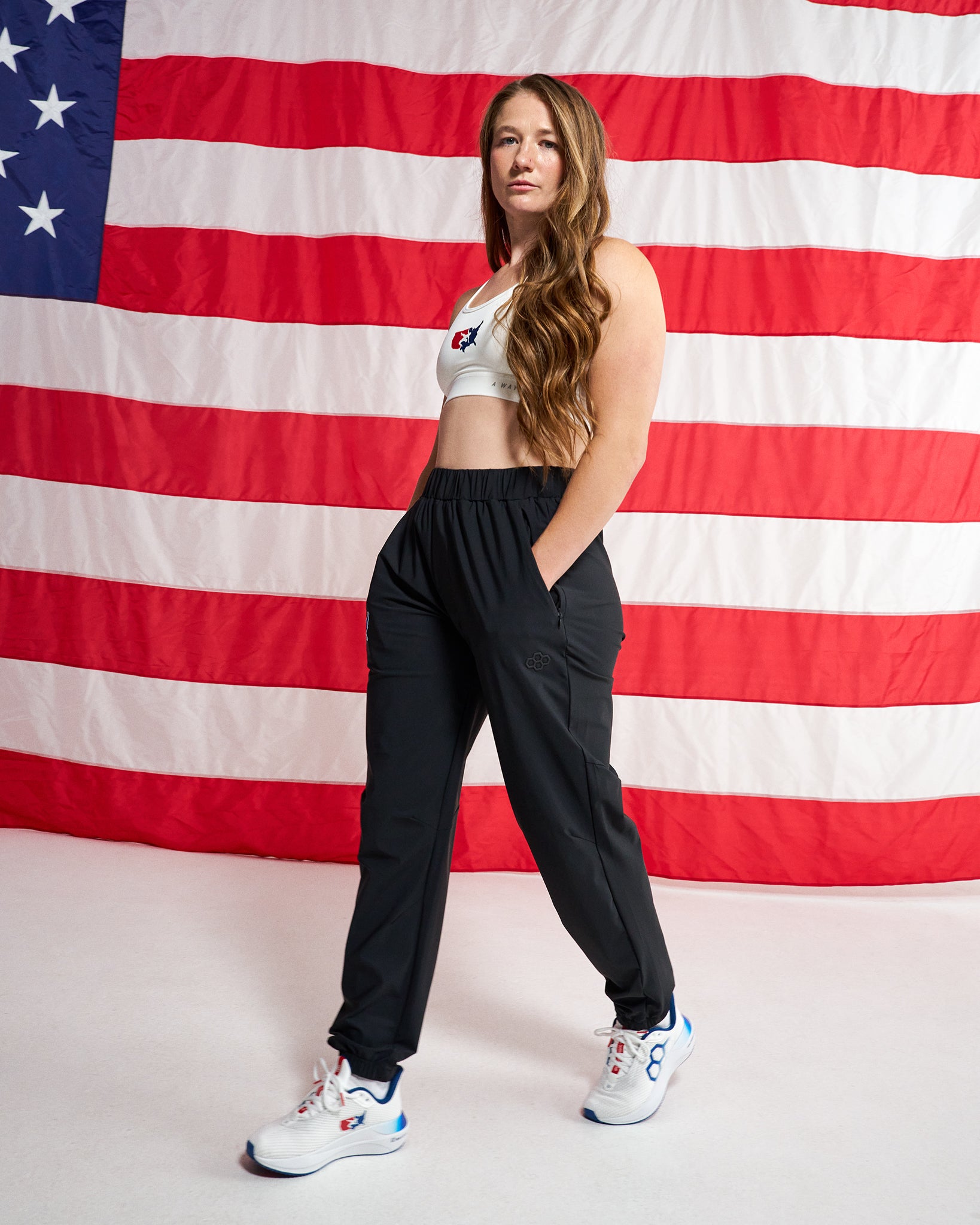 A woman modeling black athletic jogger pants with a white sports bra and white sneakers, standing in front of a large American flag backdrop.