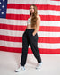 A woman modeling black athletic jogger pants with a white sports bra and white sneakers, standing in front of a large American flag backdrop.