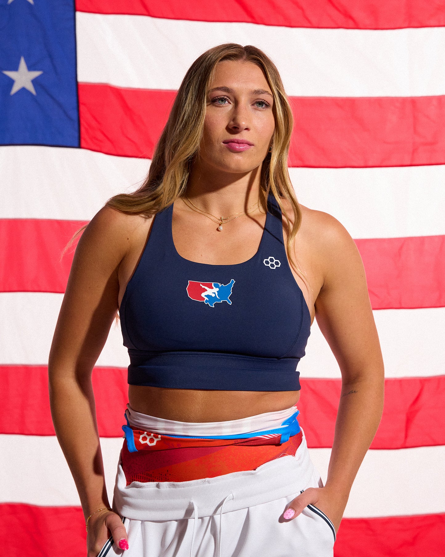 Female athlete wearing the RUDIS USA Wrestling Women's Navy Scoop Neck Padded Sports Bra stands in front of an American flag with white USA Wrestling joggers layered over colorful warm-up gear.