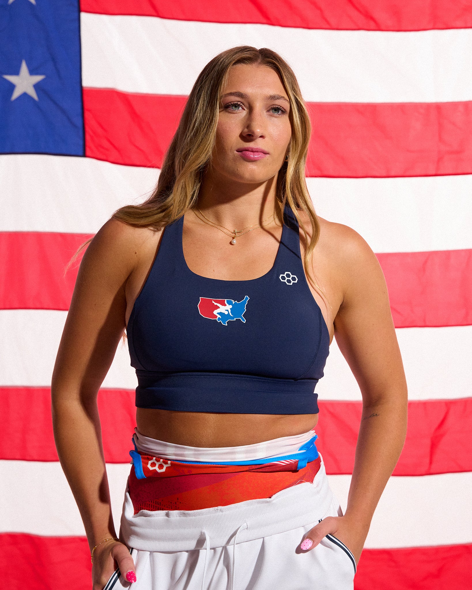 Female athlete wearing the RUDIS USA Wrestling Women's Navy Scoop Neck Padded Sports Bra stands in front of an American flag with white USA Wrestling joggers layered over colorful warm-up gear.