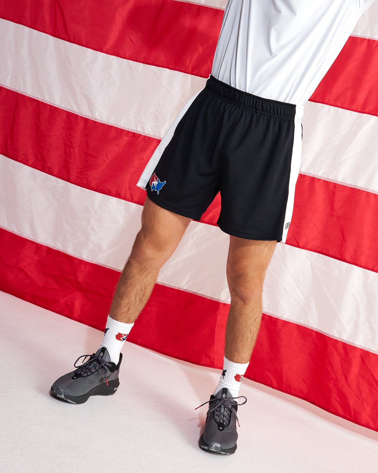 A person wearing black athletic shorts with white side stripes and a small blue fox logo, paired with white socks and dark gray running shoes, standing in front of a red and white striped flag backdrop.