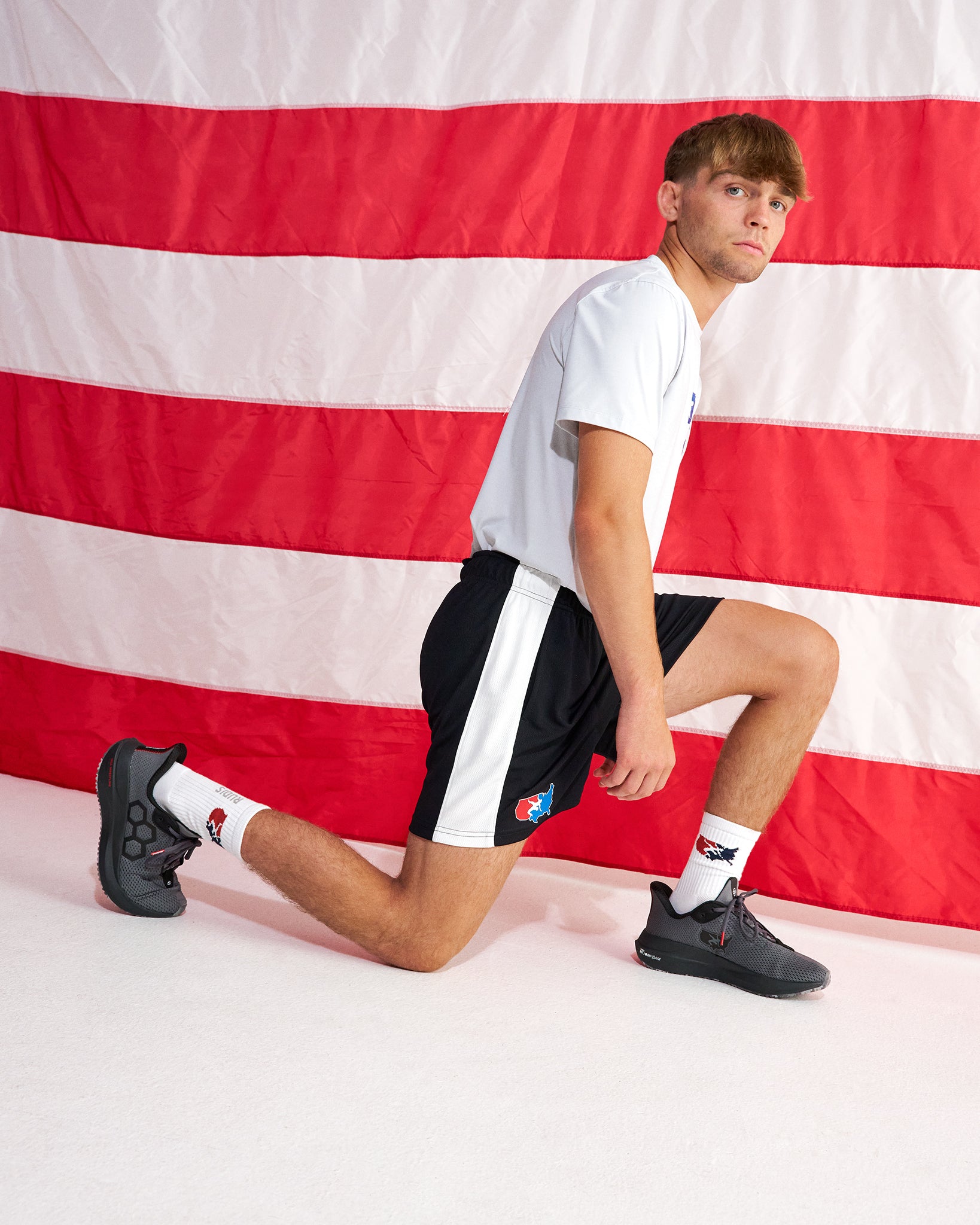 A man in a white t-shirt and black shorts with white side stripes poses in a lunge position in front of a red and white striped flag backdrop.