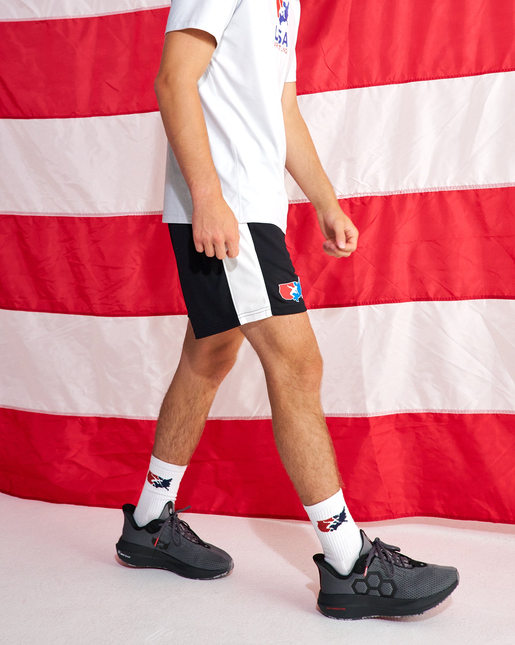 A person wearing black and white athletic shorts with a red logo, a white t-shirt, white socks, and dark gray sneakers, standing in front of a red and white striped flag backdrop.