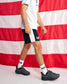 A person wearing black and white athletic shorts with a red logo, a white t-shirt, white socks, and dark gray sneakers, standing in front of a red and white striped flag backdrop.