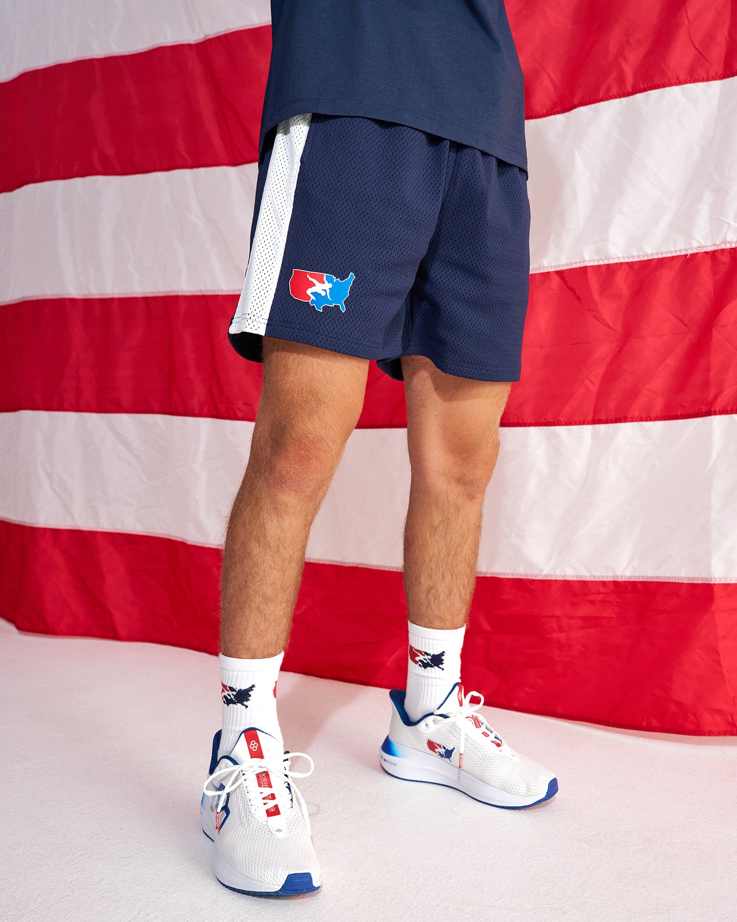 A person wearing navy athletic shorts with a white side stripe and a red, white, and blue USA map logo, paired with white socks and white sneakers, posed in front of a large American flag backdrop.