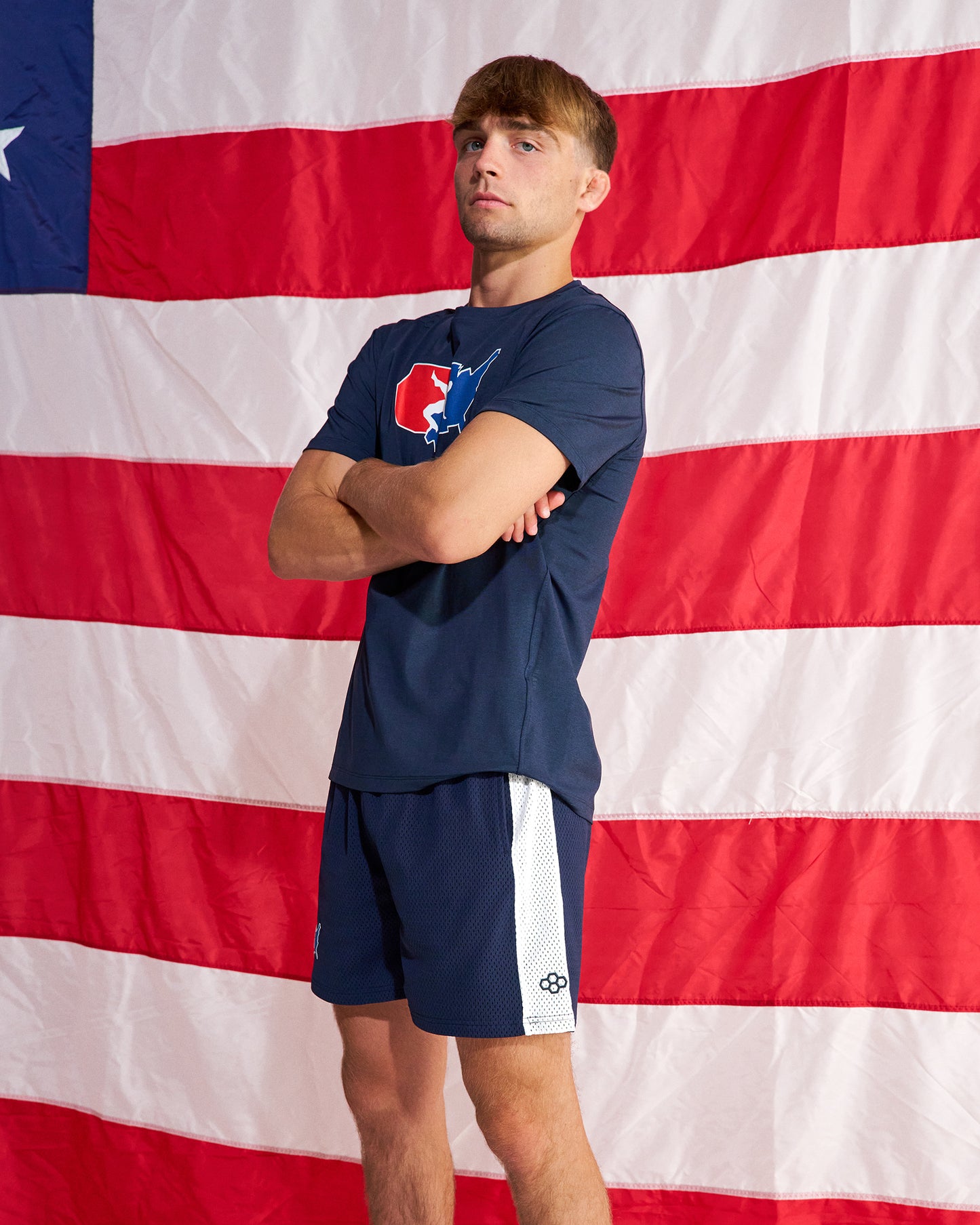 A man wearing a navy blue athletic t-shirt and matching shorts with white side stripes, standing in front of a large American flag backdrop.