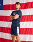 A man wearing a navy blue athletic t-shirt and matching shorts with white side stripes, standing in front of a large American flag backdrop.