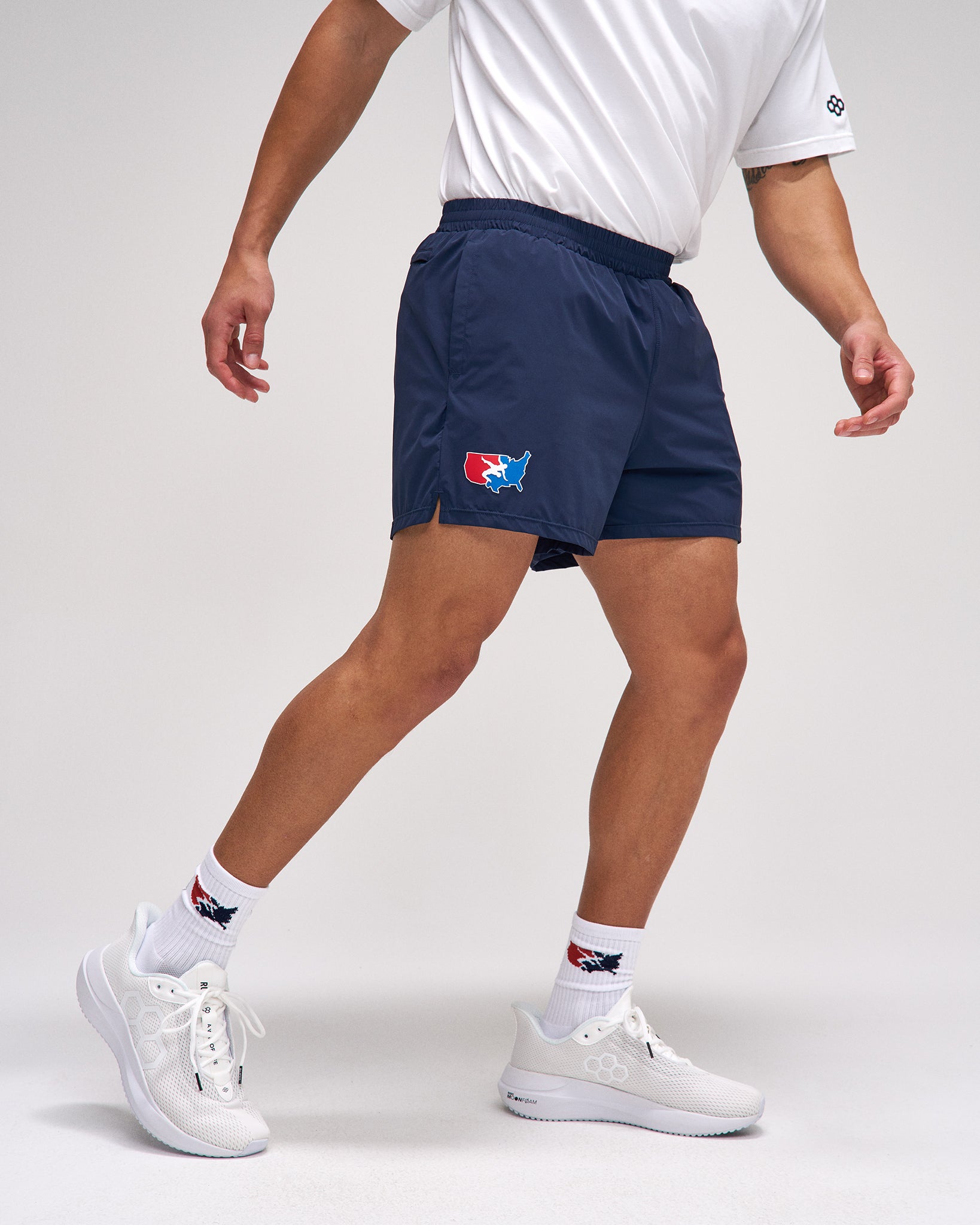 Person wearing navy athletic shorts with a logo, white shirt, and white sneakers on a light gray background