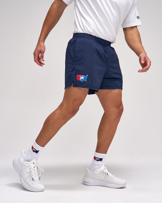 Person wearing navy athletic shorts with a logo, white shirt, and white sneakers on a light gray background