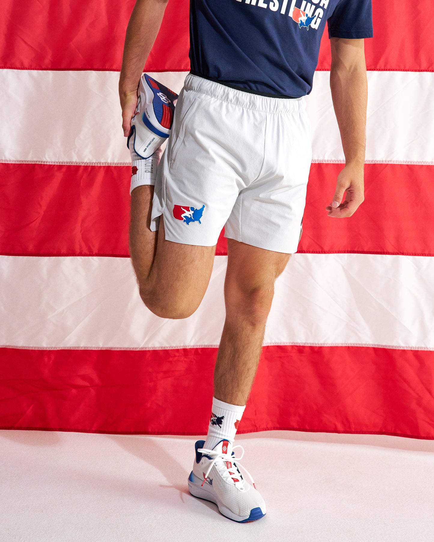A person wearing white athletic shorts with a red and blue USA Wrestling logo, a navy t-shirt, and white sneakers, stretching their leg against a red and white striped American flag background.