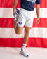 A person wearing white athletic shorts with a red and blue USA Wrestling logo, a navy t-shirt, and white sneakers, stretching their leg against a red and white striped American flag background.