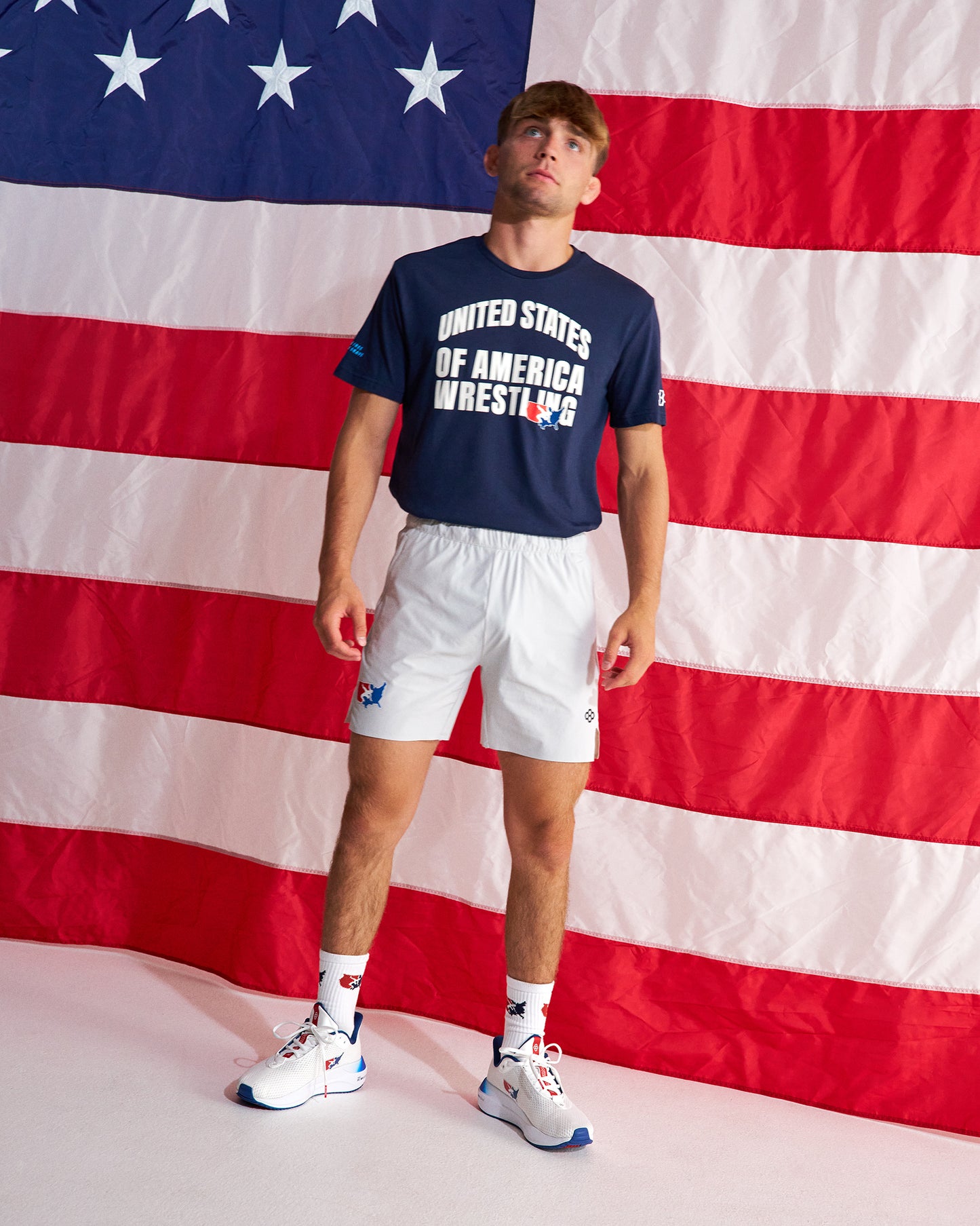 A man stands in front of a large American flag backdrop wearing a navy blue t-shirt with 'UNITED STATES OF AMERICA WRESTLING' printed in white, white athletic shorts, white socks, and white sneakers with blue accents.