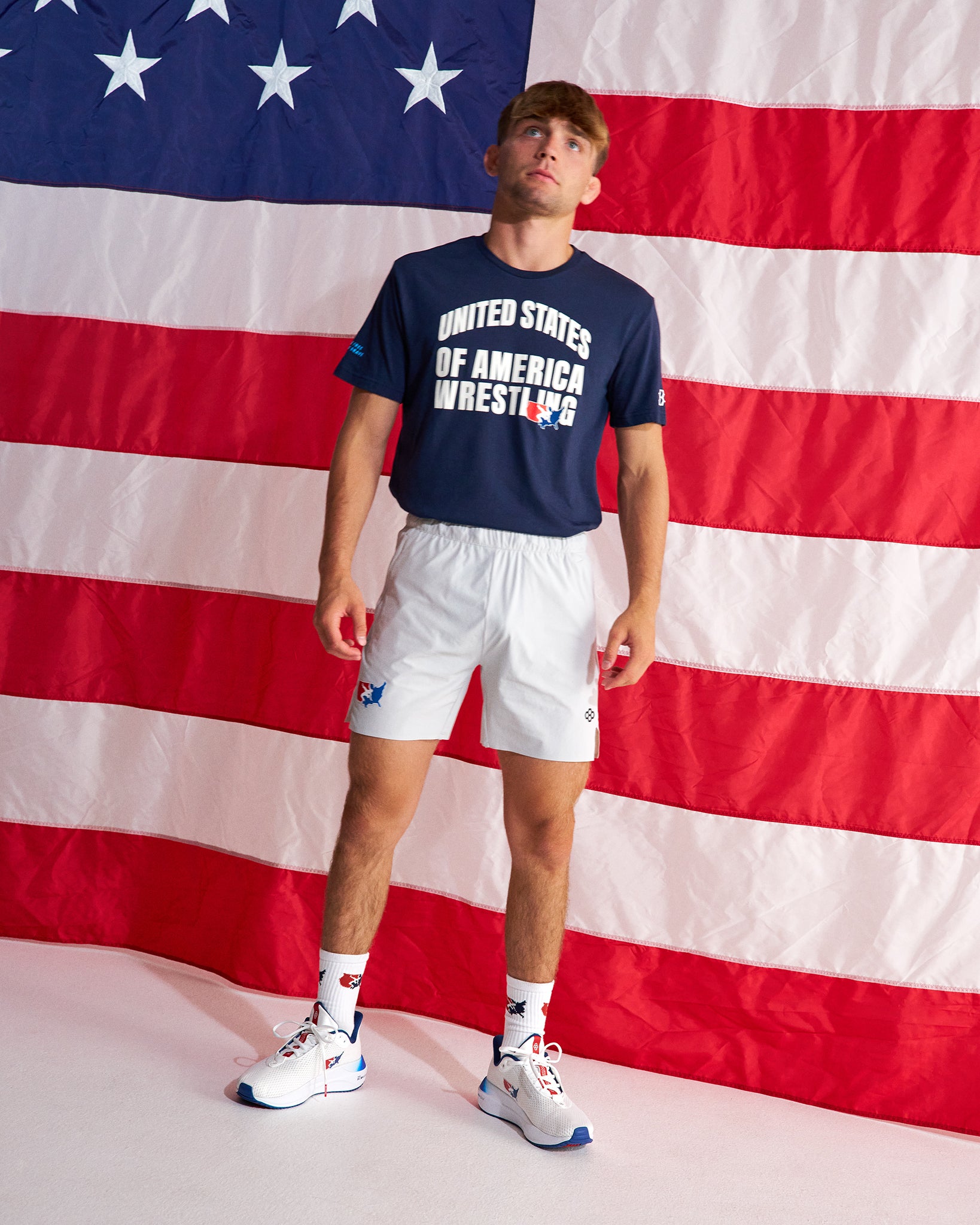 A man stands in front of a large American flag backdrop wearing a navy blue t-shirt with 'UNITED STATES OF AMERICA WRESTLING' printed in white, white athletic shorts, white socks, and white sneakers with blue accents.