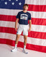 A man stands in front of a large American flag backdrop wearing a navy blue t-shirt with 'UNITED STATES OF AMERICA WRESTLING' printed in white, white athletic shorts, white socks, and white sneakers with blue accents.