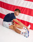 A man in a navy blue athletic t-shirt, light gray shorts, white socks, and blue-and-white sneakers sits on a white floor in front of a large red and white striped flag, stretching his leg.