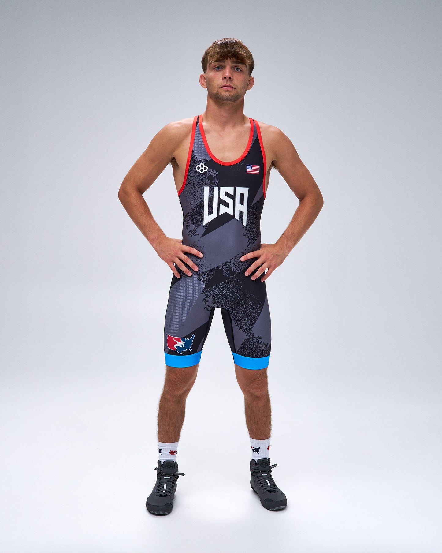 A male athlete stands with hands on hips wearing a USA wrestling singlet. The singlet is dark gray with a subtle pattern, featuring red trim, white 'USA' lettering on the chest, a small American flag, and bright blue accents on the shorts.
