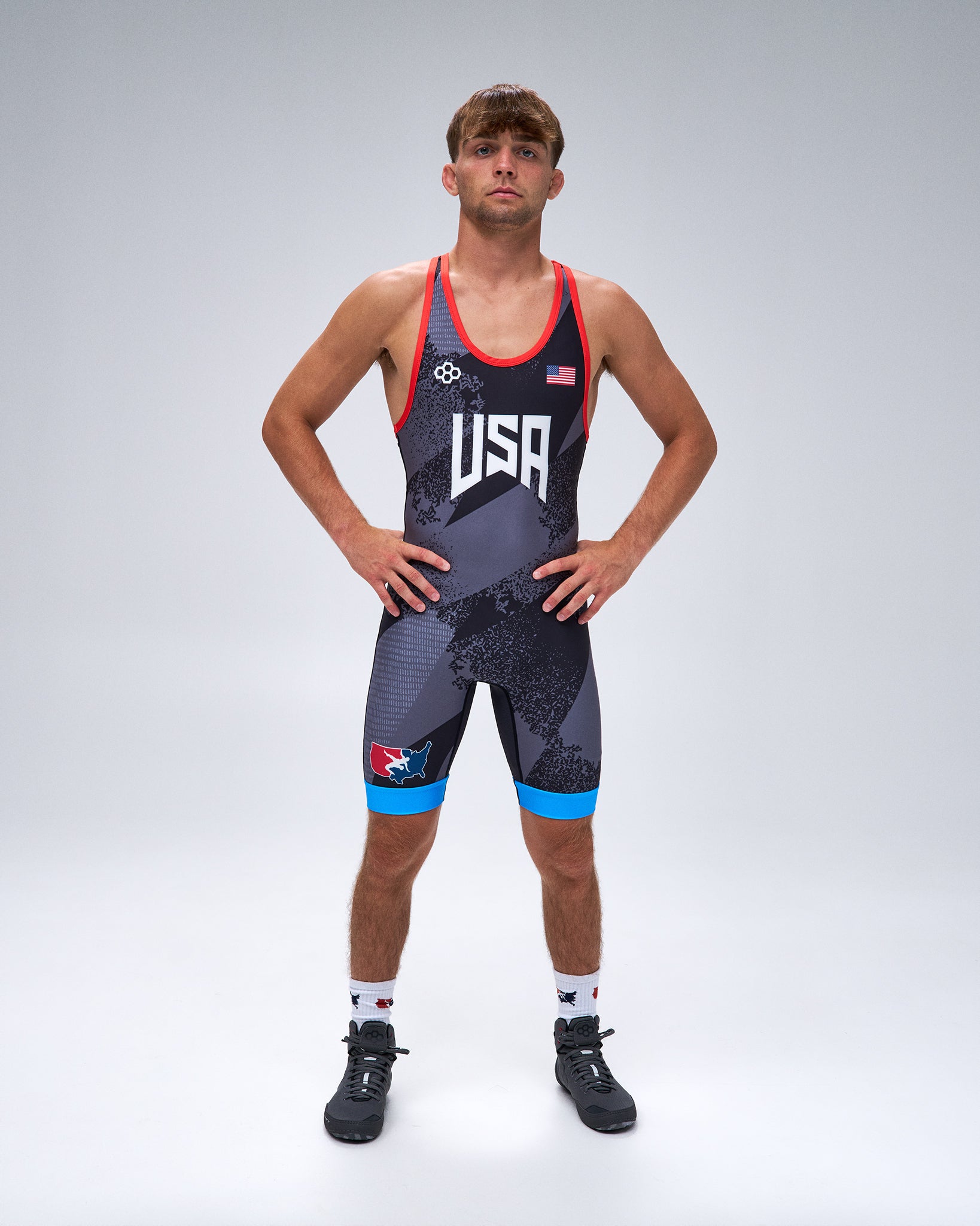 A male athlete stands with hands on hips wearing a USA wrestling singlet. The singlet is dark gray with a subtle pattern, featuring red trim, white 'USA' lettering on the chest, a small American flag, and bright blue accents on the shorts.