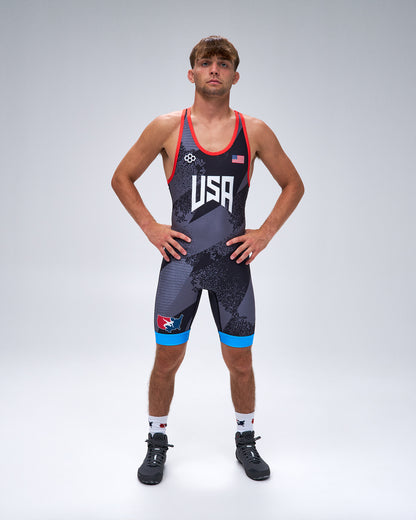 A male athlete stands with hands on hips wearing a USA wrestling singlet. The singlet is dark gray with a subtle pattern, featuring red trim, white 'USA' lettering on the chest, a small American flag, and bright blue accents on the shorts.