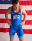A man wearing a blue USA Wrestling singlet with orange trim, standing in front of an American flag backdrop.