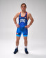 A man stands in a studio wearing a blue USA wrestling singlet with orange trim, white 'USA' lettering, and small American flag and logo details. He is also wearing dark wrestling shoes and white socks against a light gray background.