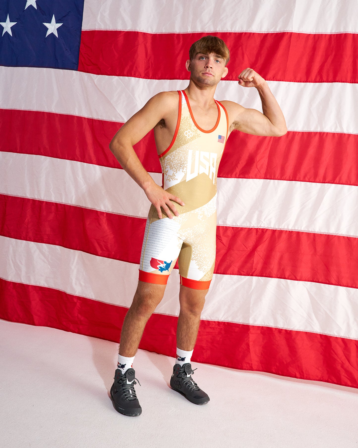 A man wearing a beige and white wrestling singlet with red trim poses in front of a large American flag backdrop, flexing his arm. He is wearing black athletic shoes and white socks.