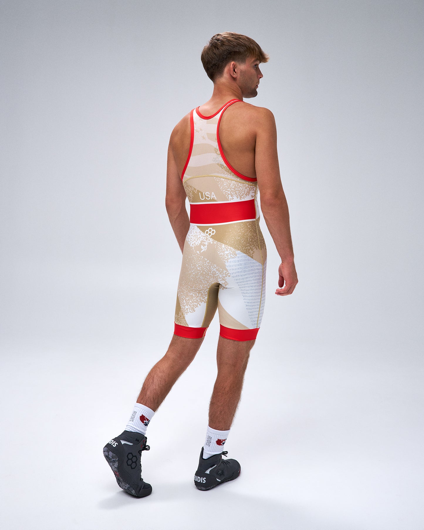 A man wearing a sleeveless Wrestling singlet with a gold, white, and red pattern, shown from a rear three-quarter view against a light gray studio background.