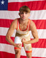 A young man wearing a USA wrestling singlet poses in front of a large American flag. The singlet is beige with red trim and features 'USA' printed across the chest.