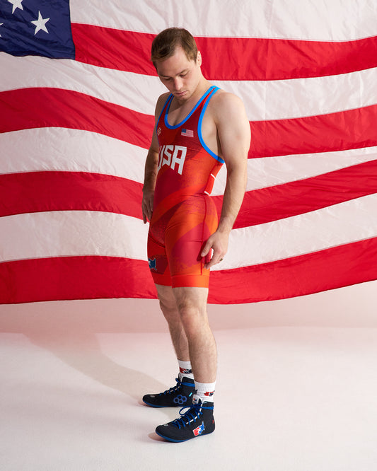 A man wearing a red and orange USA wrestling singlet with blue trim, standing in front of a large American flag backdrop.