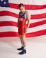 A man wearing a red and orange USA wrestling singlet with blue trim, standing in front of a large American flag backdrop.