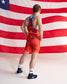 A male athlete shown from behind wearing a red and orange USA wrestling singlet with blue trim, standing in front of a large American flag backdrop.