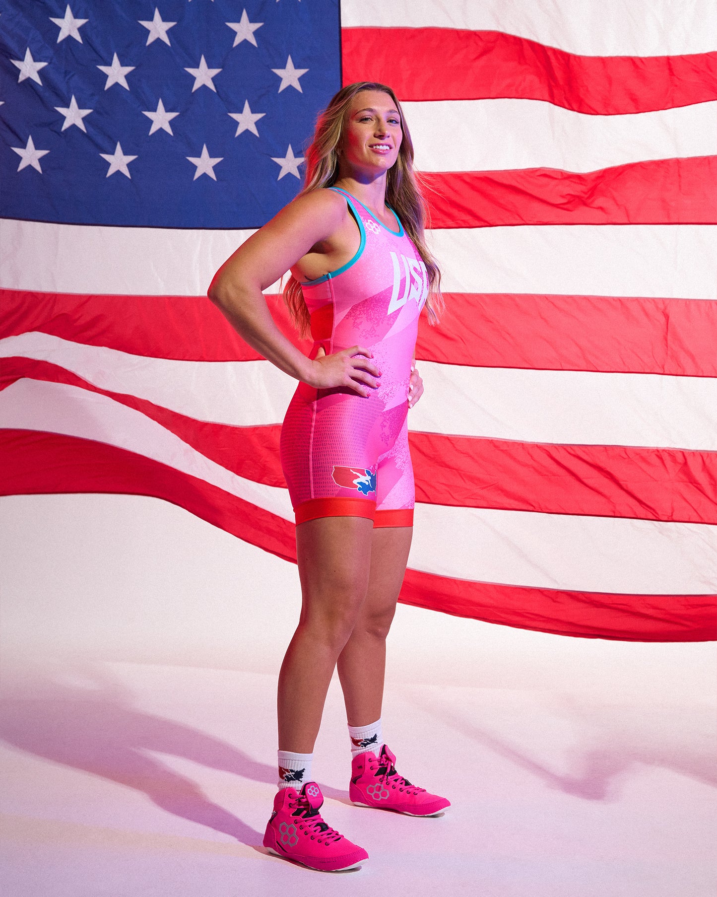 A woman stands in front of a large American flag backdrop wearing a bright pink wrestling singlet with blue trim, white lettering, and matching pink athletic shoes.
