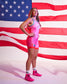 A woman stands in front of a large American flag backdrop wearing a bright pink wrestling singlet with blue trim, white lettering, and matching pink athletic shoes.